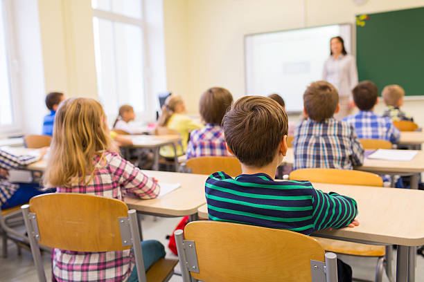 education, elementary school, learning and people concept - group of school kids sitting and listening to teacher in classroom from back