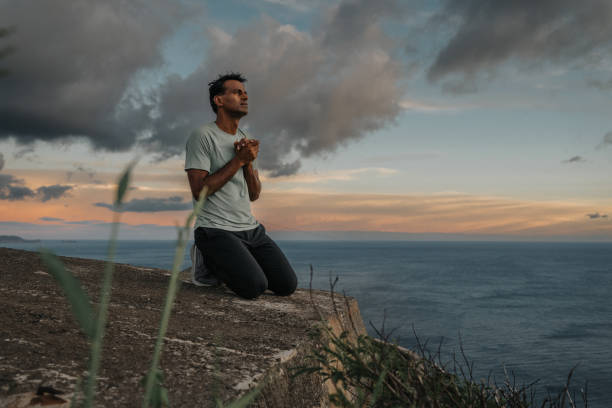 Pursue divine presence.- A Christian man of Indian descent kneels on the ground at a scenic viewpoint overlooking the Pacific Ocean at sunrise and folds his hands in prayer. Hope, spirituality and gratitude concepts.